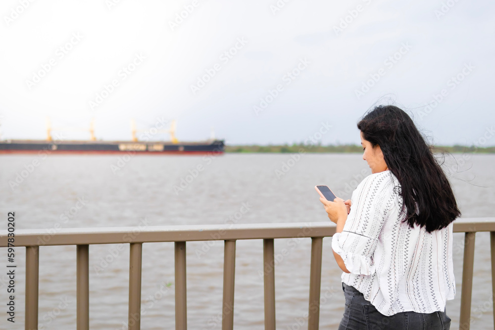 Young person in Barranquilla using technology while chatting on their cellphone, with an impressive cargo ship in the background. 
