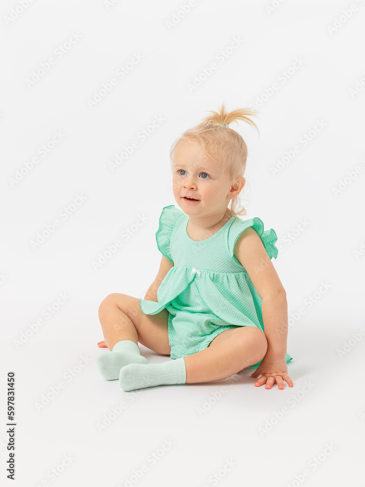 A 2-year-old toddler girl attentively looks at something with interest. Sitting on a white background in a green dress. A 2-year-old toddler girl attentively looks at something with interest. Sitting on a white background in a green dress.