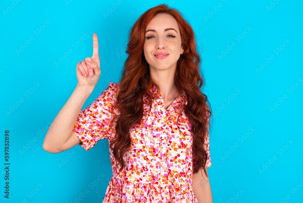 Caucasian redhead woman wearing floral dress over blue background showing and pointing up with fingers number one while smiling confident and happy. Caucasian redhead woman wearing floral dress over blue background showing and pointing up with fingers number one while smiling confident and happy.