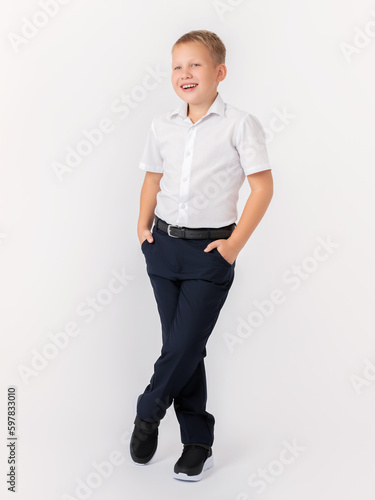 A cute 10-year-old blonde teenager stands cross-legged in a classic white shirt and dark trousers with a belt on a white background. Hands in pockets. A glance to the side. Layout, advertising.