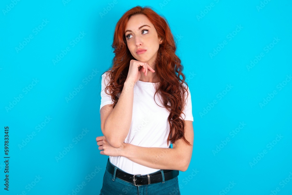 Face expressions and emotions. Thoughtful young redhead woman wearing white T-shirt over blue background holding hand under his head, having doubtful look. Face expressions and emotions. Thoughtful young redhead woman wearing white T-shirt over blue background holding hand under his head, having doubtful look.