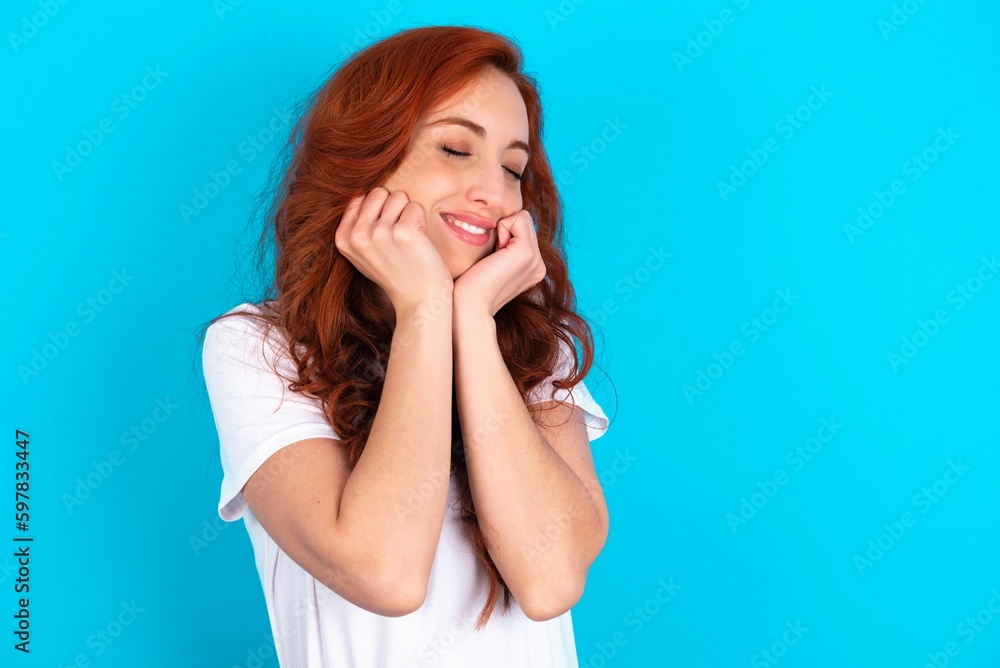 Portrait of young redhead woman wearing white T-shirt over blue background being overwhelmed, expressing excitement and happiness with closed eyes and hands near face. Portrait of young redhead woman wearing white T-shirt over blue background being overwhelmed, expressing excitement and happiness with closed eyes and hands near face.
