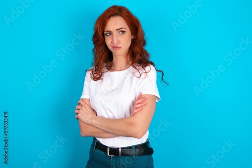 Photo Displeased young redhead woman wearing white T-shirt over blue background with bad attitude, arms crossed looking sideways
