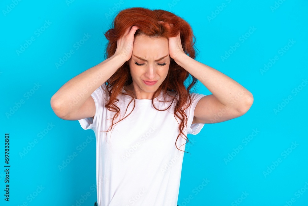 young redhead woman wearing white T-shirt over blue background holding head with hands, suffering from severe headache, pressing fingers to temples young redhead woman wearing white T-shirt over blue background holding head with hands, suffering from severe headache, pressing fingers to temples