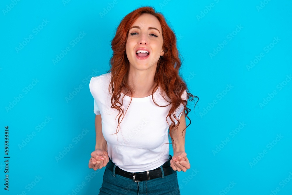 young redhead woman wearing white T-shirt over blue background raising fists up screaming with joy being happy to achieve goals. young redhead woman wearing white T-shirt over blue background raising fists up screaming with joy being happy to achieve goals.