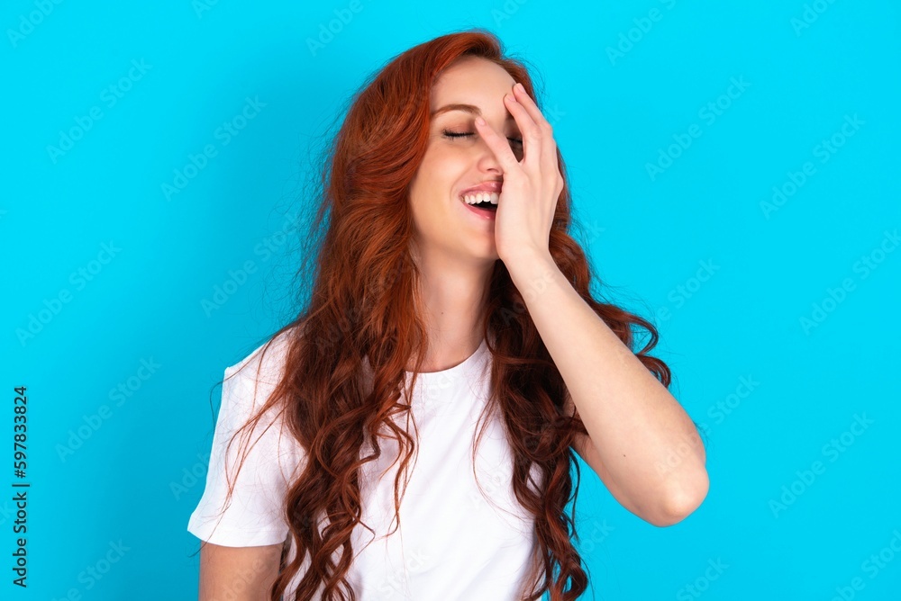 Charismatic carefree joyful young redhead woman wearing white T-shirt over blue background likes laugh out loud not hiding emotions giggling hear funny hilarious joke chuckling facepalm. Charismatic carefree joyful young redhead woman wearing white T-shirt over blue background likes laugh out loud not hiding emotions giggling hear funny hilarious joke chuckling facepalm.
