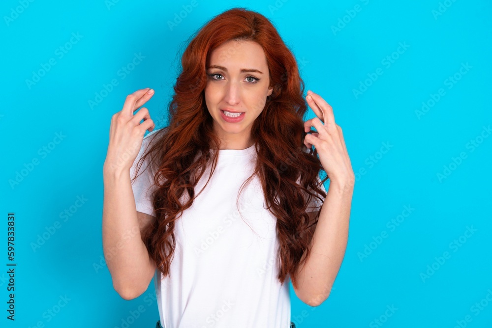 Fototapeta premium young redhead woman wearing white T-shirt over blue background holding fingers crossed with worried expression hoping boss didn't noticed mistakes at work.