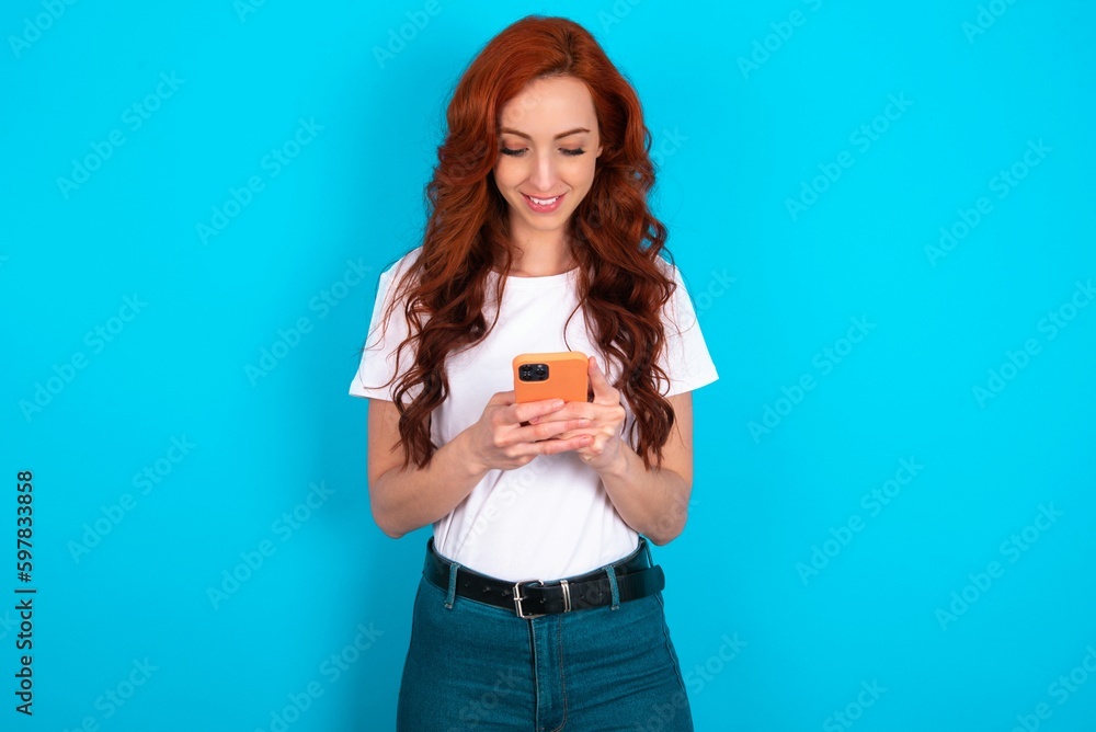Smiling young redhead woman wearing white T-shirt over blue background using cell phone, messaging, being happy to text with friends, looking at smartphone. Modern technologies and communication. Smiling young redhead woman wearing white T-shirt over blue background using cell phone, messaging, being happy to text with friends, looking at smartphone. Modern technologies and communication.