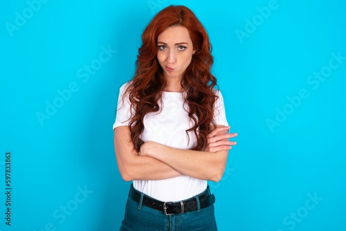 Picture of angry young redhead woman wearing white T-shirt over blue background crossing arms. Looking at camera with disappointed expression.