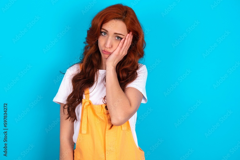 Sad lonely Young redhead woman wearing orange overall over blue background touches cheek with hand bites lower lip and gazes with displeasure. Bad emotions Sad lonely Young redhead woman wearing orange overall over blue background touches cheek with hand bites lower lip and gazes with displeasure. Bad emotions