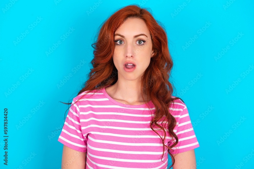 Shocked young redhead woman wearing striped T-shirt over blue background stares bugged eyes keeps mouth opened has surprised expression. Omg concept Shocked young redhead woman wearing striped T-shirt over blue background stares bugged eyes keeps mouth opened has surprised expression. Omg concept