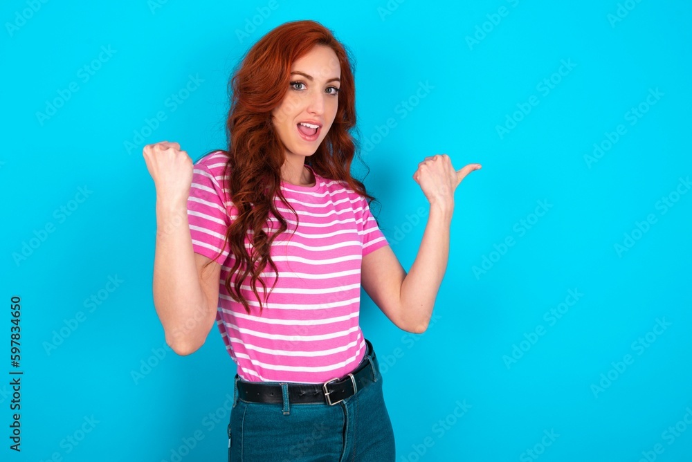 Hooray cool young redhead woman wearing striped T-shirt over blue background point back empty space hand fist Hooray cool young redhead woman wearing striped T-shirt over blue background point back empty space hand fist