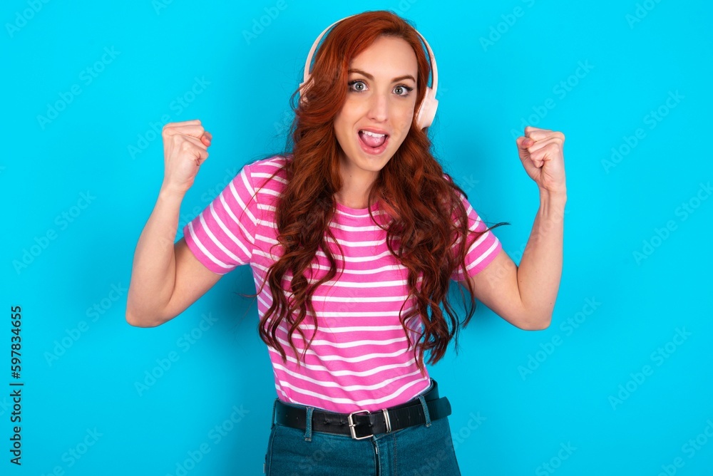 Fototapeta premium Emotional young redhead woman wearing striped T-shirt over blue background exclaims loudly feels like winner raises clenched fists keeps mouth opened wears stereo headphones on ears makes yes gesture