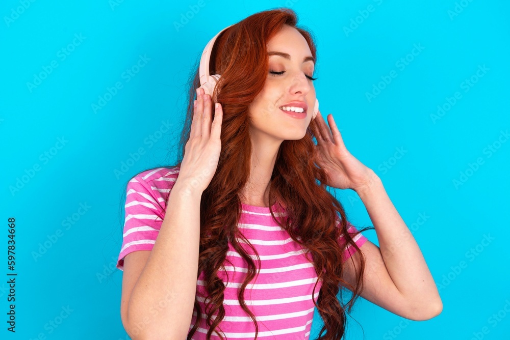 young redhead woman wearing striped T-shirt over blue background with headphones on head, listens to music, enjoying favourite song with closed eyes, holding hands on headset. young redhead woman wearing striped T-shirt over blue background with headphones on head, listens to music, enjoying favourite song with closed eyes, holding hands on headset.