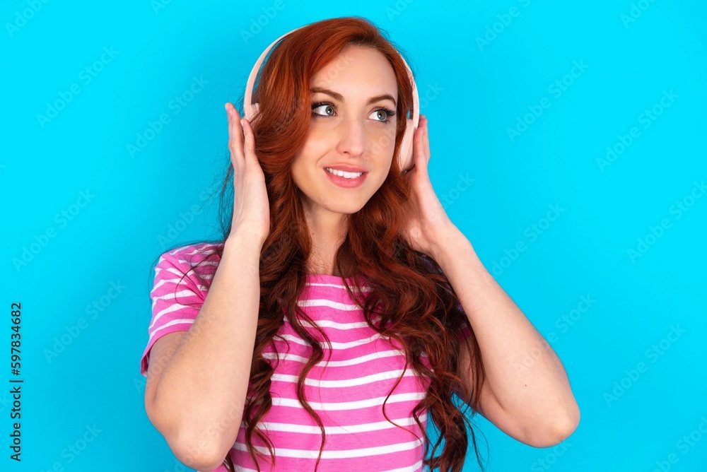 young redhead woman wearing striped T-shirt over blue background wears stereo headphones listens music concentrated aside. People hobby lifestyle concept young redhead woman wearing striped T-shirt over blue background wears stereo headphones listens music concentrated aside. People hobby lifestyle concept