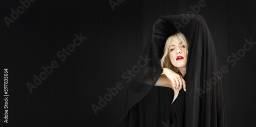 Wide banner portrait of blonde fashion beauty model woman, with red mouth and black fabric, cloth with hair electrostatic charged, sticking to cape, against black background