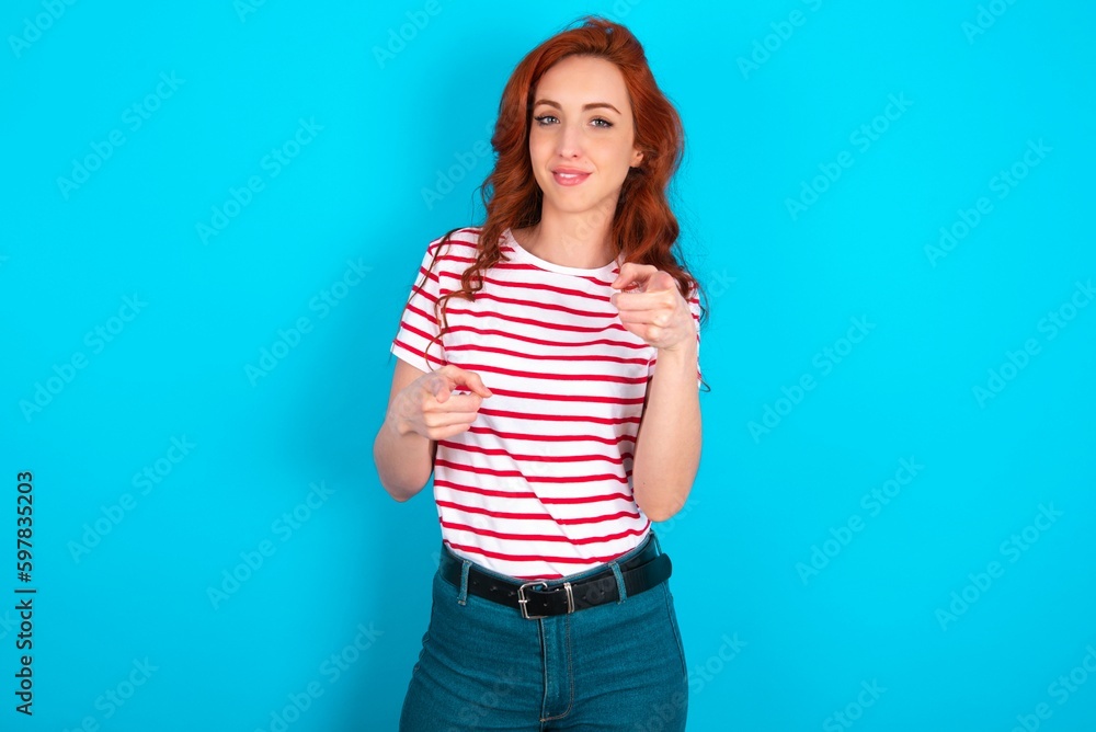 young redhead woman wearing striped T-shirt over blue background pointing fingers to camera with happy and funny face. Good energy and vibes. young redhead woman wearing striped T-shirt over blue background pointing fingers to camera with happy and funny face. Good energy and vibes.