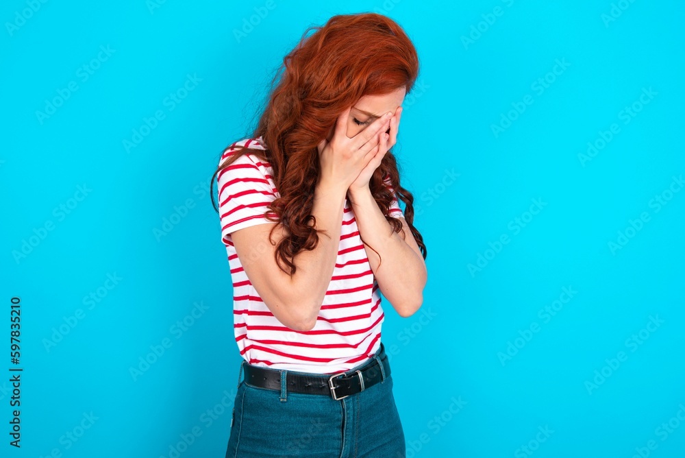 Sad young redhead woman wearing striped T-shirt over blue background crying covering her face with her hands. Sad young redhead woman wearing striped T-shirt over blue background crying covering her face with her hands.