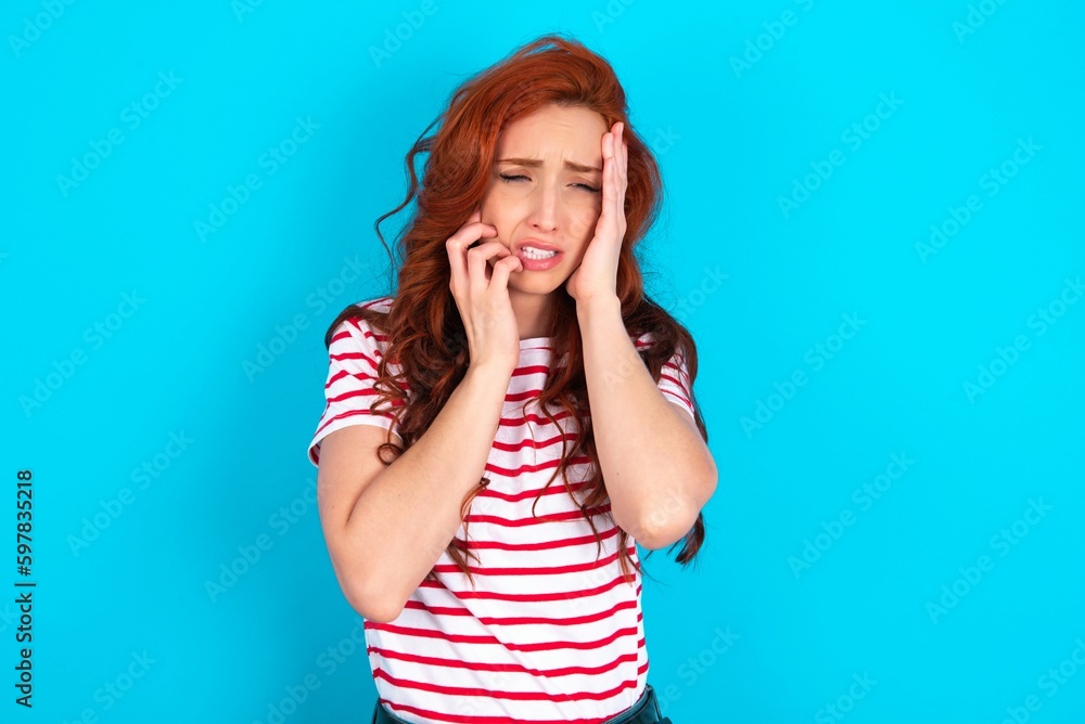 Doleful desperate crying young redhead woman wearing striped T-shirt over blue background , looks stressfully, frowns face, feels lonely and anxious Doleful desperate crying young redhead woman wearing striped T-shirt over blue background , looks stressfully, frowns face, feels lonely and anxious