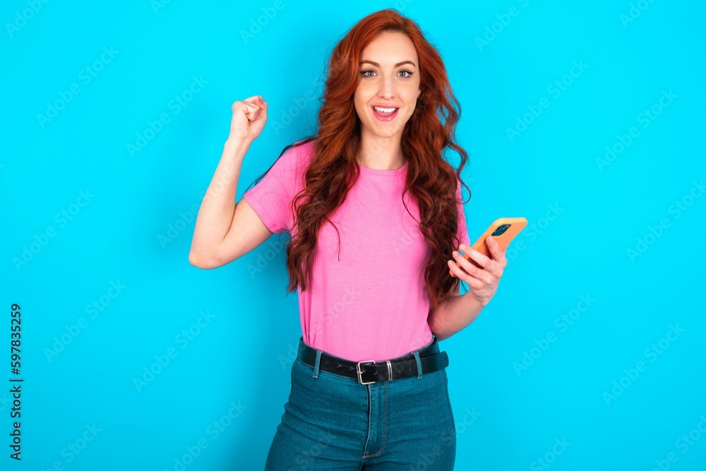 Portrait of young redhead woman wearing pink T-shirt over blue background holding in hands cell celebrating Portrait of young redhead woman wearing pink T-shirt over blue background holding in hands cell celebrating