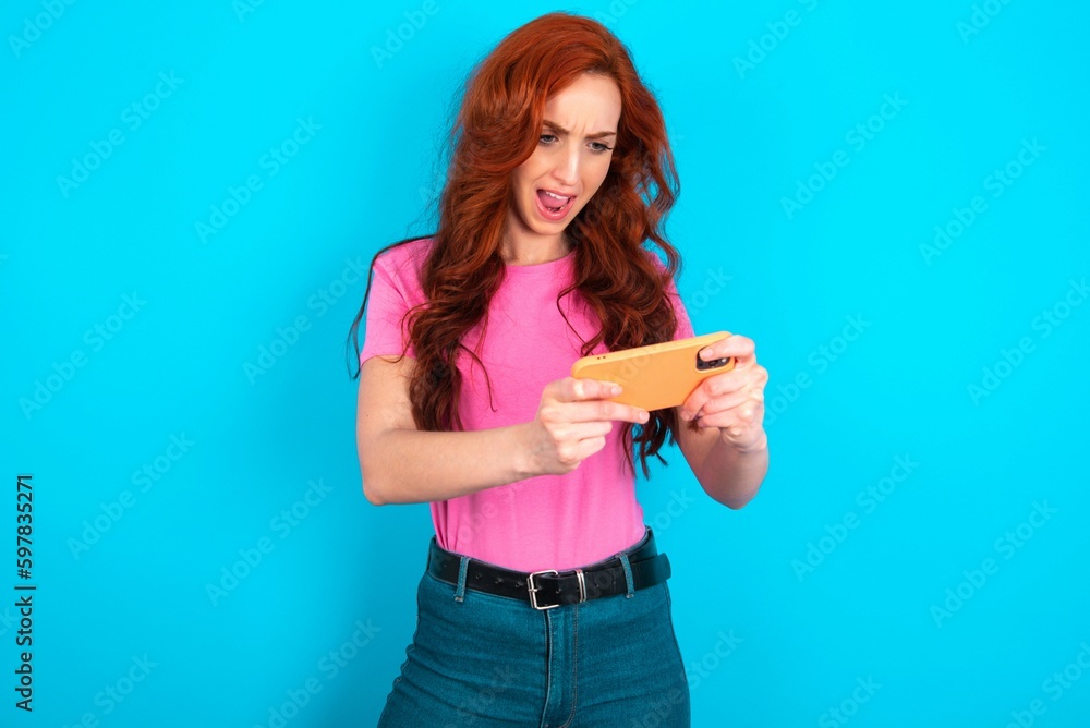 Portrait of an excited young redhead woman wearing pink T-shirt over blue background playing games on mobile phone. Portrait of an excited young redhead woman wearing pink T-shirt over blue background playing games on mobile phone.