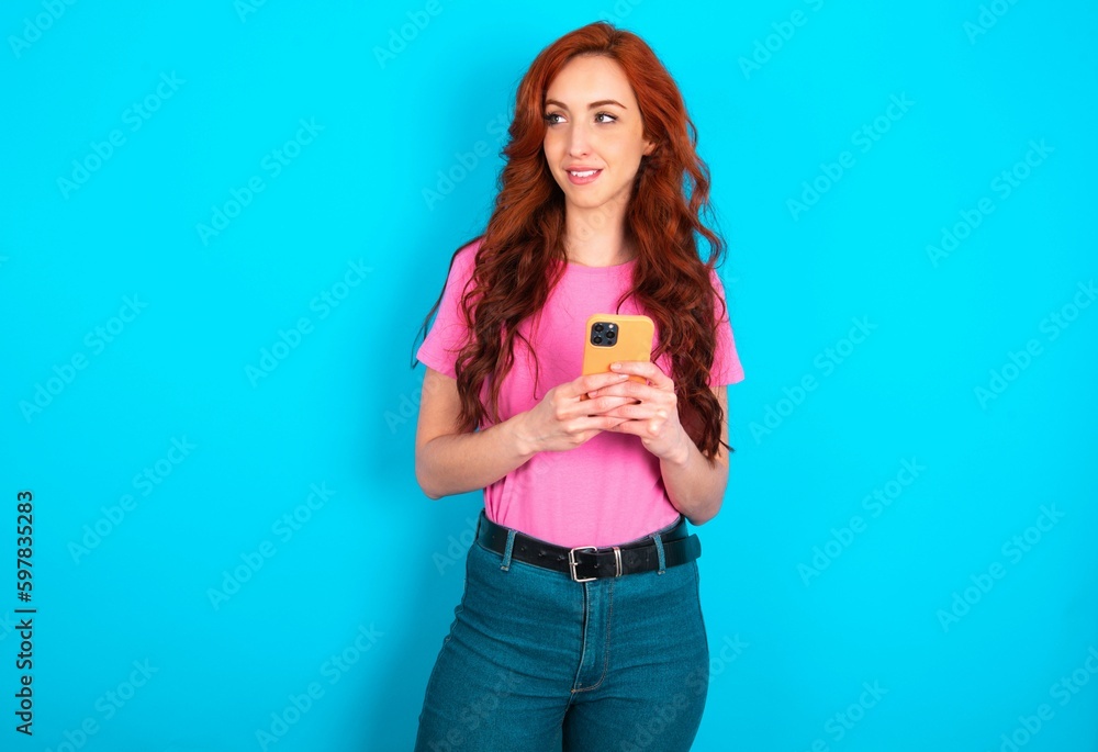 young redhead woman wearing pink T-shirt over blue background hold telephone hands read good youth news look empty space advert young redhead woman wearing pink T-shirt over blue background hold telephone hands read good youth news look empty space advert