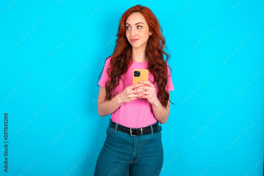 young redhead woman wearing pink T-shirt over blue background holds telephone hands reads good youth news look empty space advert young redhead woman wearing pink T-shirt over blue background holds telephone hands reads good youth news look empty space advert