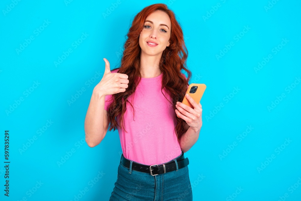 Portrait of young redhead woman wearing pink T-shirt over blue background using and texting with smartphone happy with big smile doing ok sign, thumb up with fingers, excellent sign Portrait of young redhead woman wearing pink T-shirt over blue background using and texting with smartphone happy with big smile doing ok sign, thumb up with fingers, excellent sign