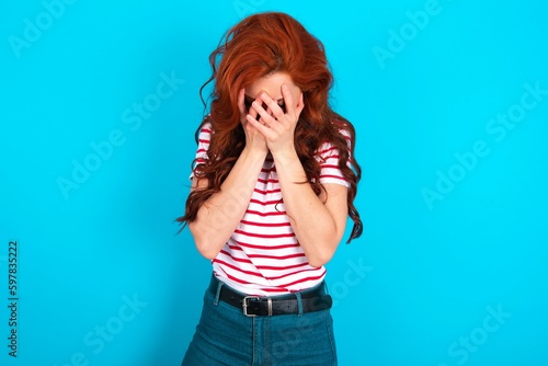 young redhead woman wearing striped T-shirt over blue background covering her face with her hands, being devastated and crying. Sad concept