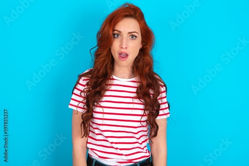 Oh my God. Surprised young redhead woman wearing striped T-shirt over blue background stares at camera with shocked expression exclaims with unexpectedness,