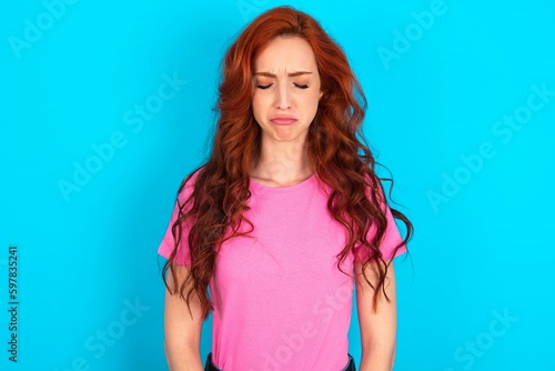 Fototapeta Dismal gloomy rejected young redhead woman wearing pink T-shirt over blue backgr