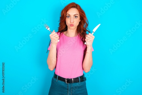 hungry young redhead woman wearing pink T-shirt over blue background holding in hand fork knife want tasty yummy pizza pie