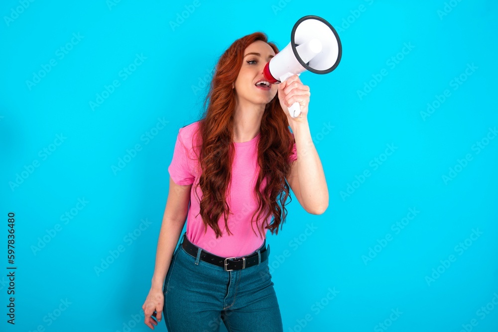 young redhead woman wearing pink T-shirt over blue background Through Megaphone with Available Copy Space young redhead woman wearing pink T-shirt over blue background Through Megaphone with Available Copy Space