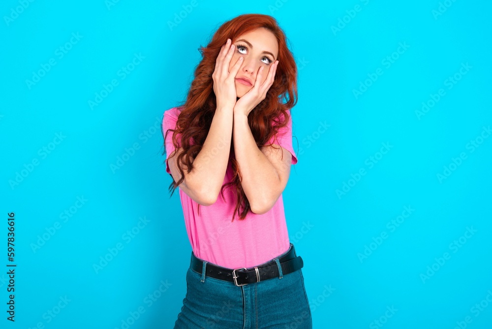 young redhead woman wearing pink T-shirt over blue background keeps hands on cheeks has bored displeased expression. Stressed hopeless model young redhead woman wearing pink T-shirt over blue background keeps hands on cheeks has bored displeased expression. Stressed hopeless model
