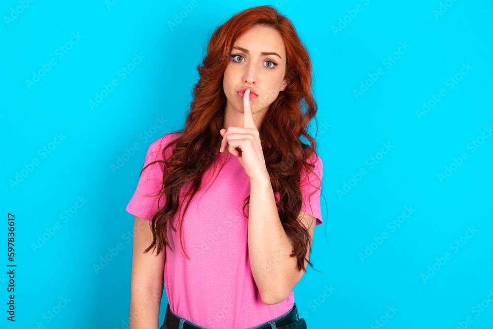 Surprised young redhead woman wearing pink T-shirt over blue background makes silence gesture, keeps finger over lips and looks mysterious at camera Surprised young redhead woman wearing pink T-shirt over blue background makes silence gesture, keeps finger over lips and looks mysterious at camera