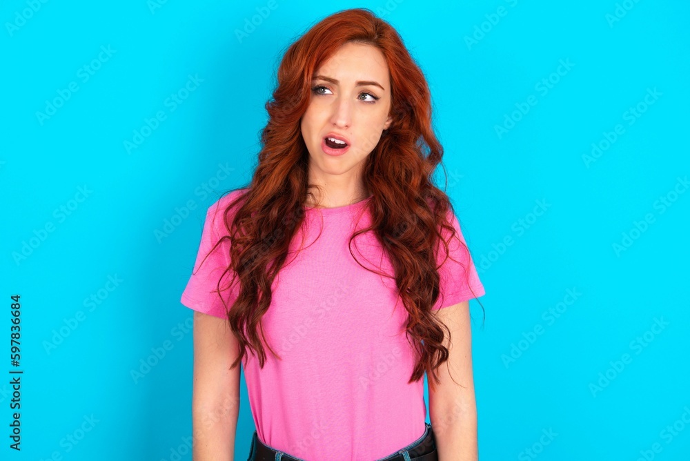 young redhead woman wearing pink T-shirt over blue background yawns with opened mouth stands. Daily morning routine young redhead woman wearing pink T-shirt over blue background yawns with opened mouth stands. Daily morning routine