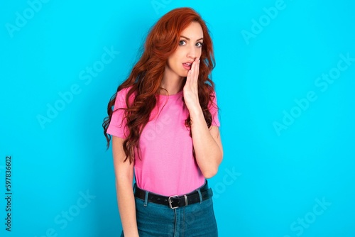 young redhead woman wearing pink T-shirt over blue background covers mouth and looks with wonder at camera, cannot believe unexpected rumors.