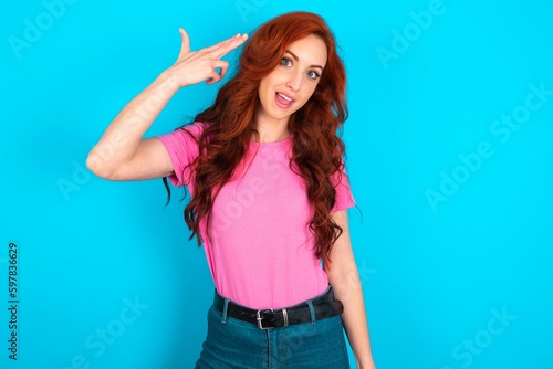 Vászonkép young redhead woman wearing pink T-shirt over blue background foolishness around shoots in temple with fingers makes suicide gesture
