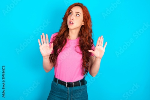 young redhead woman wearing pink T-shirt over blue background keeps palms forward and looks with fright above on ceiling tries to defense herself from invisible danger opens mouth.