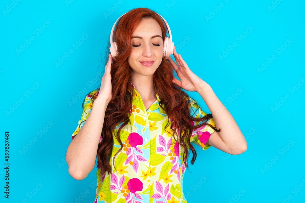 Pleased Young redhead woman wearing colorful shirt over blue background enjoys listening pleasant melody keeps hands on stereo headphones closes eyes. Spending free time with music Pleased Young redhead woman wearing colorful shirt over blue background enjoys listening pleasant melody keeps hands on stereo headphones closes eyes. Spending free time with music