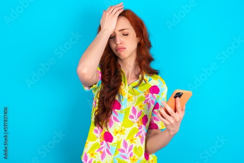 Upset depressed Young redhead woman wearing colorful shirt over blue background makes face palm as forgot about something important holds mobile phone expresses sorrow and regret blames