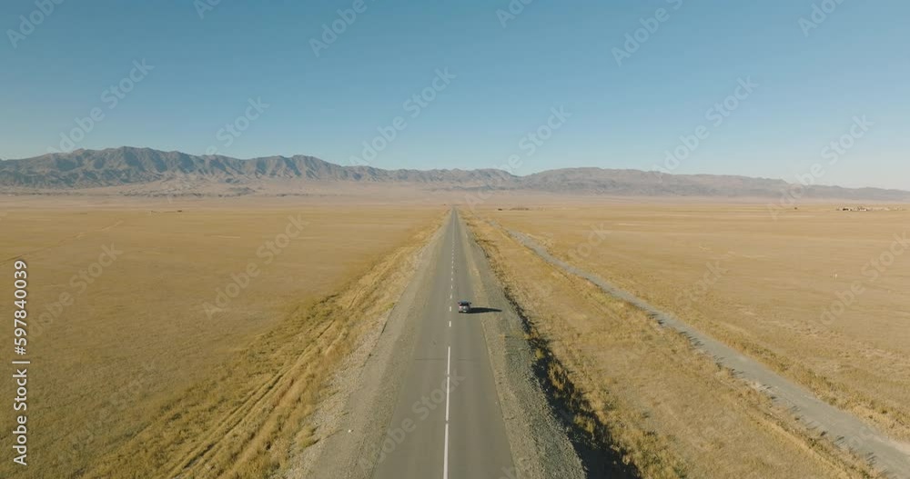 One gray crossover SUV car driving alone, travel on empty straight asphalt road freeway through empty arid steppe plain at summer sunny day. Aerial drone wide into the distance view. One gray crossover SUV car driving alone, travel on empty straight asphalt road freeway through empty arid steppe plain at summer sunny day. Aerial drone wide into the distance view.