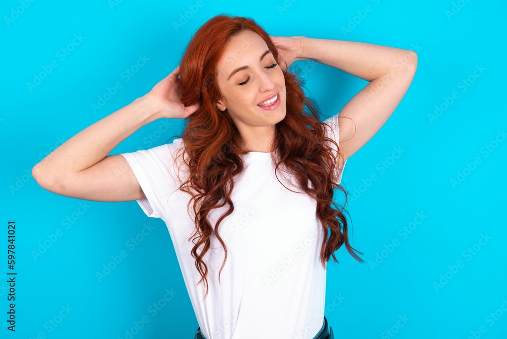 young redhead woman wearing white T-shirt over blue background relaxing and stretching, arms and hands behind head and neck smiling happy