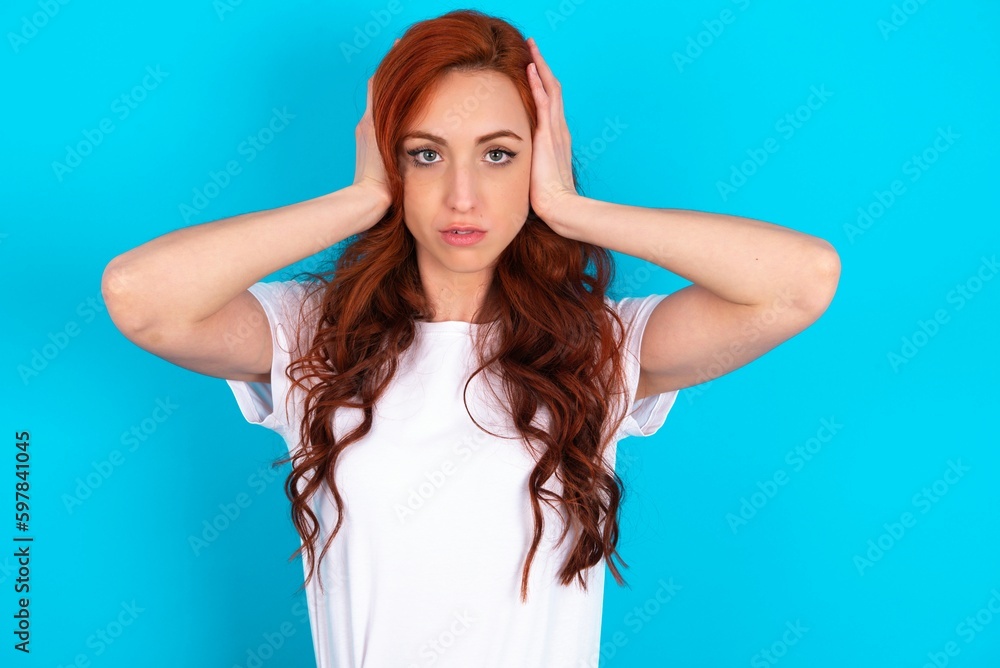 Frustrated young redhead woman wearing white T-shirt over blue background plugging ears with hands does not wanting to listen hard rock, noise or loud music.