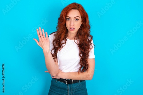 Photo Studio shot of frustrated young redhead woman wearing white T-shirt over blue background  gesturing with raised palm, frowning, being displeased and confused with dumb question