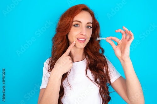 young redhead woman wearing white T-shirt over blue background holding an invisible aligner and pointing to her perfect straight teeth. Dental healthcare and confidence concept.