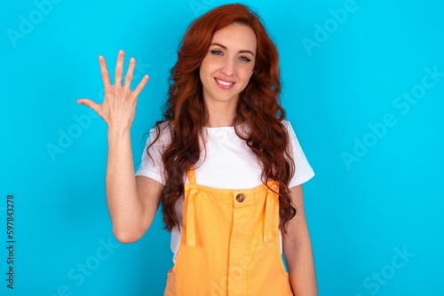 Young redhead woman wearing orange overall over blue background smiling and looking friendly, showing number five or fifth with hand forward, counting down