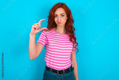 Displeased young redhead woman wearing striped T-shirt over blue background shapes little hand sign demonstrates something not very big. Body language concept.