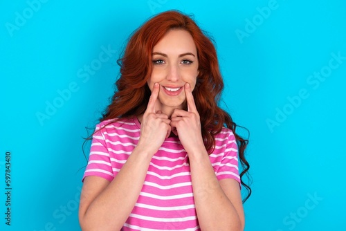 Happy young redhead woman wearing striped T-shirt over blue background with toothy smile, keeps index fingers near mouth, fingers pointing and forcing cheerful smile