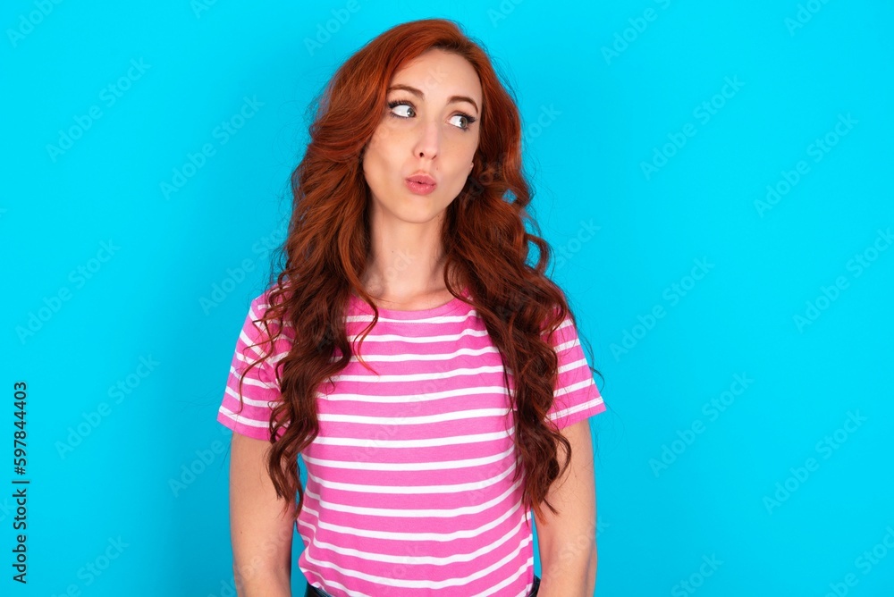 Shocked young redhead woman wearing striped T-shirt over blue background look empty space with open mouth screaming: Oh My God! I can't believe this. Shocked young redhead woman wearing striped T-shirt over blue background look empty space with open mouth screaming: Oh My God! I can't believe this.